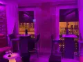 Ambiente moderno y acogedor para comer en Coruña. Mesas altas, taburetes y sofás rojos bajo luces violetas.