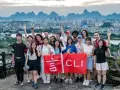 Grupo de jóvenes sonríe y saluda, sosteniendo una bandera roja con caracteres chinos y "CLI". Un paisaje urbano y montañoso de China al fondo, ideal para viajar a China por libre.