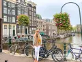 Mujer sonriente posa junto a bicicletas en un canal de Ámsterdam, con edificios históricos y flores colgantes. ¡Excelentes consejos para viajar a Ámsterdam!