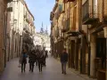 Gente pasea por una calle porticada, un lugar ideal para descubrir qué ver en Burgo de Osma. Edificios históricos y torres se alzan al fondo.