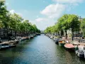 Canal de Ámsterdam con barcos, árboles y edificios bajo un cielo azul con nubes.