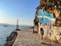 Una mujer posa junto a una roca con la bandera griega pintada. Un faro y un velero adornan la costa, ideal para saber qué ver en Grecia en una semana.