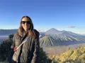 Mujer sonríe con gafas de sol frente a un paisaje volcánico en su ruta por Indonesia.