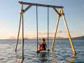 Mujer en columpio sobre el mar, ideal para saber donde viajar en febrero. Paisaje idílico con veleros y montañas al atardecer.