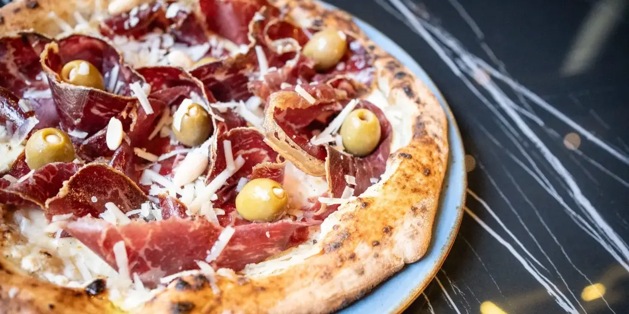 Pizza con jamón serrano, aceitunas y queso rallado, perfecta para saber donde comer en Menorca.