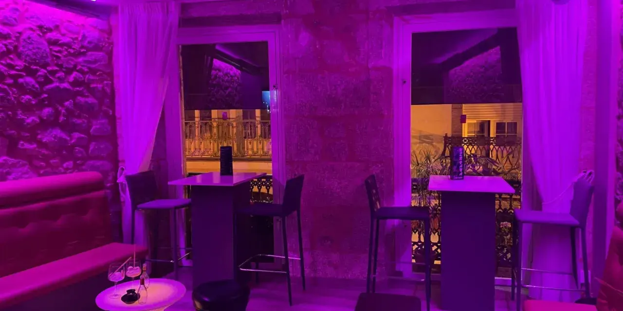 Ambiente moderno y acogedor para comer en Coruña. Mesas altas, taburetes y sofás rojos bajo luces violetas.