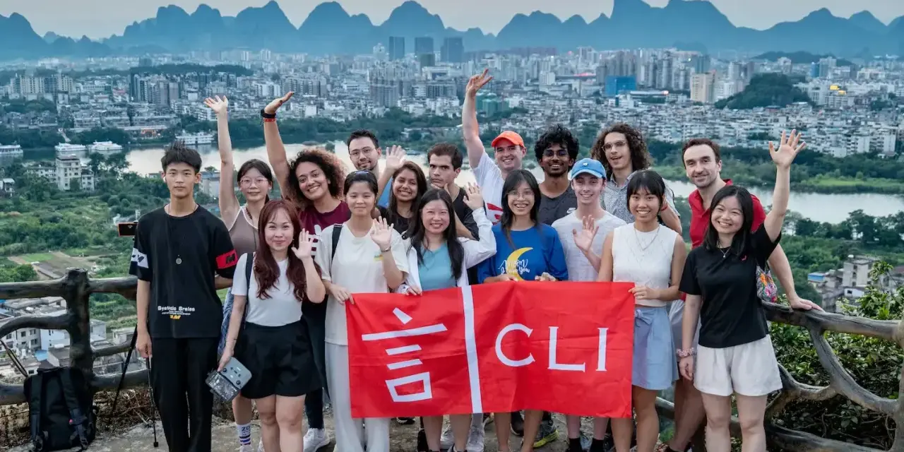 Grupo de jóvenes sonríe y saluda, sosteniendo una bandera roja con caracteres chinos y "CLI". Un paisaje urbano y montañoso de China al fondo, ideal para viajar a China por libre.