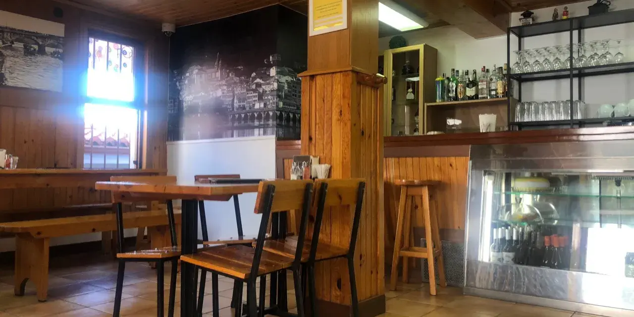 Mesas de madera y sillas listas para comer en Pontedeume. Barra con botellas y vasos, y un mural de la ciudad.