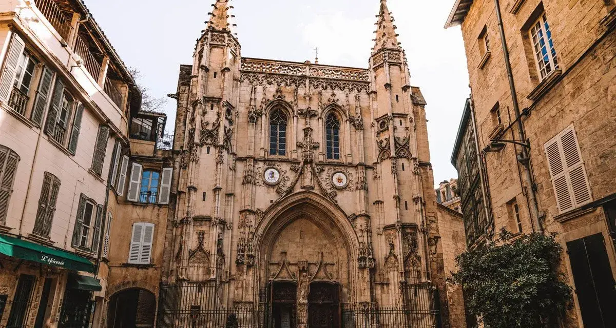 Fachada de una iglesia gótica con detalles ornamentados y dos torres. A los lados, edificios antiguos de piedra y fachadas de color claro. Un lugar que ver en Avignon.