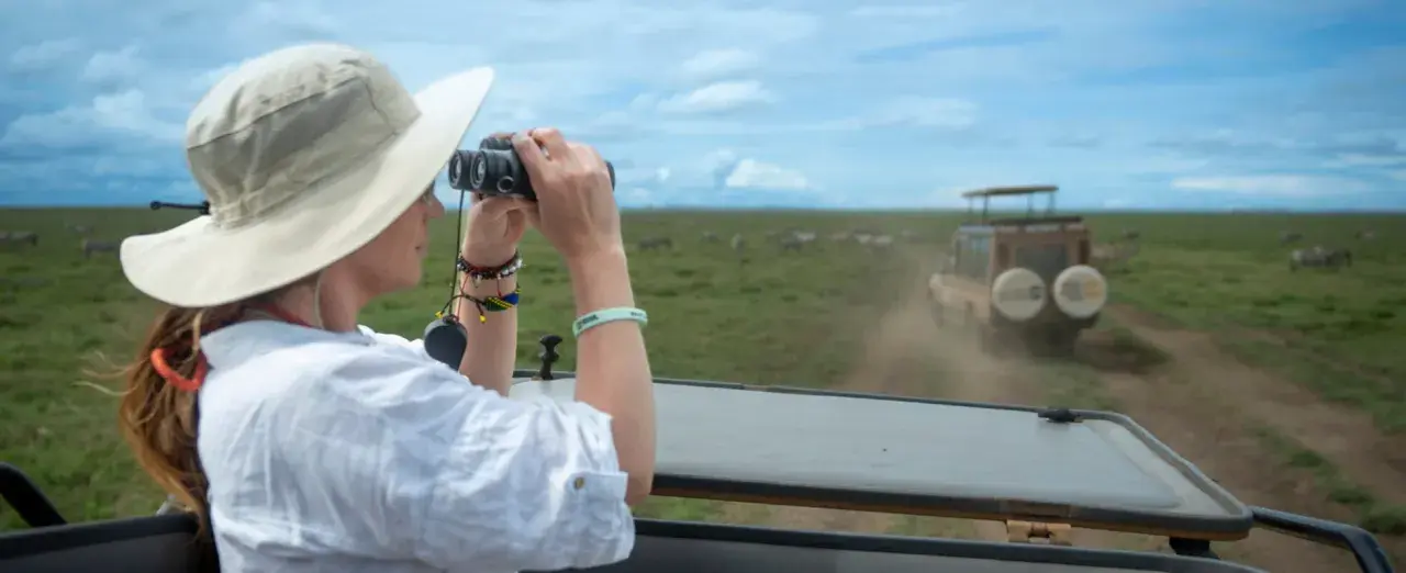 Mujer con sombrero y binoculares observa cebras en la sabana, disfrutando de su viaje a Tanzania.