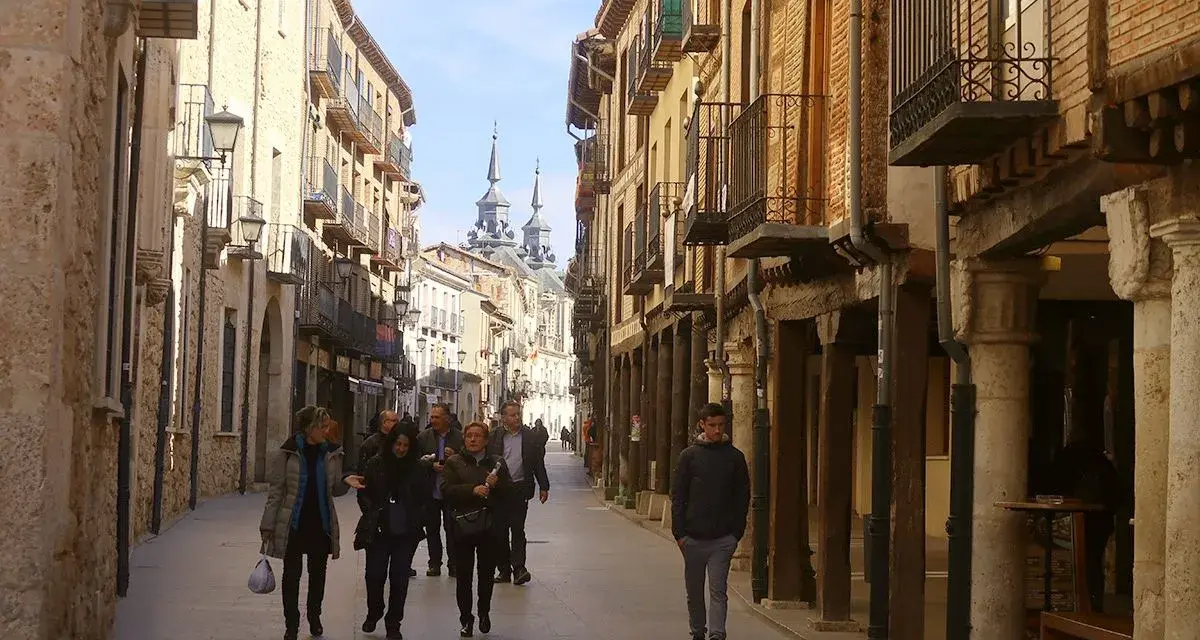 Gente pasea por una calle porticada, un lugar ideal para descubrir qué ver en Burgo de Osma. Edificios históricos y torres se alzan al fondo.