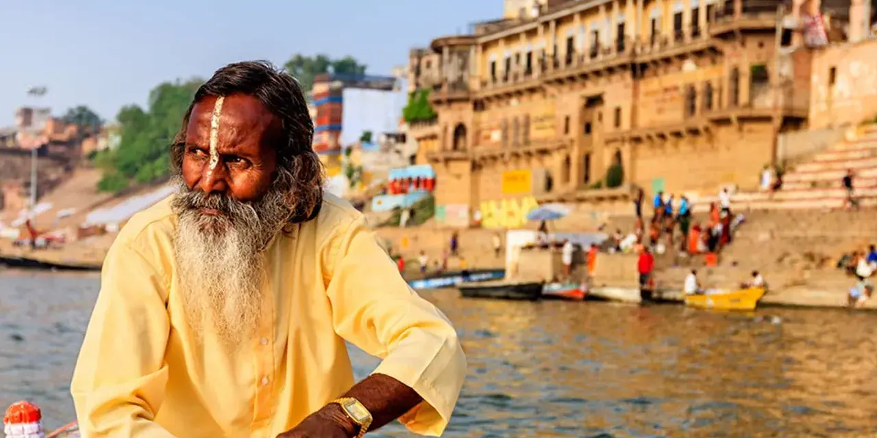 Un hombre con barba y marca en la frente navega en el Ganges, una escena típica al viajar a la India.