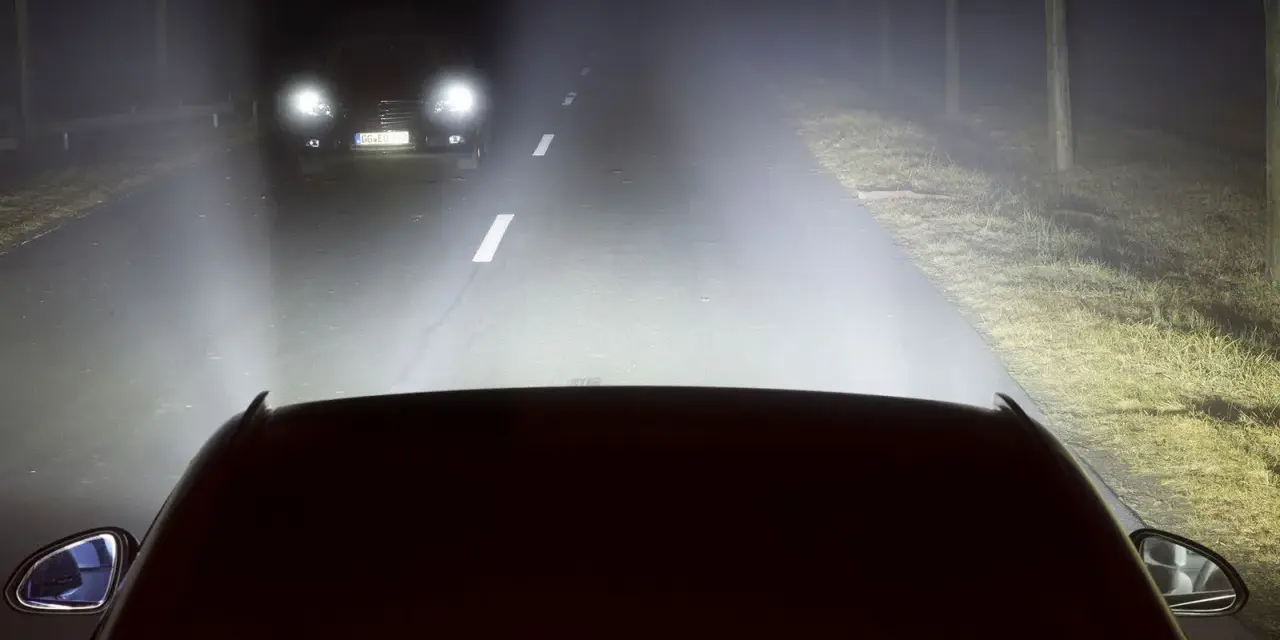 Luces de carretera o largas iluminan la noche, revelando un coche adelante y el camino.