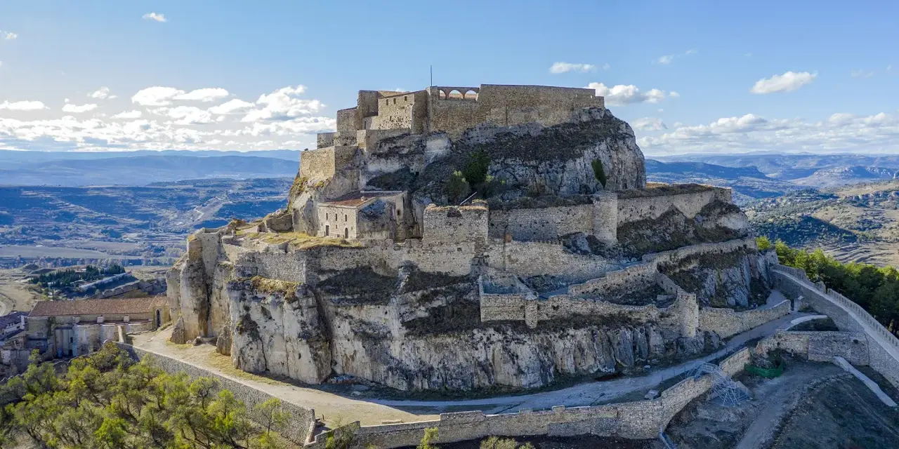 Castillo de Morella sobre un peñón rocoso, con murallas y edificios de piedra bajo un cielo azul con nubes.