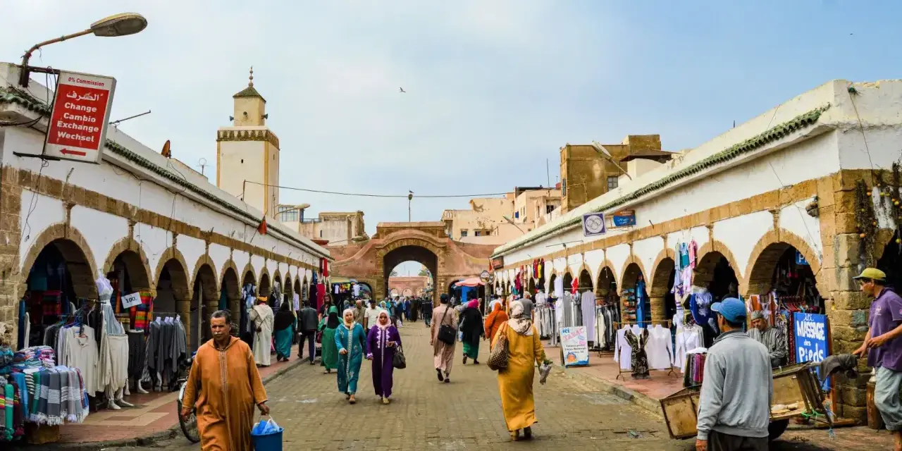 Mercado vibrante en Essaouira que ver: gente comprando ropa y artesanías bajo arcos blancos, con una mezquita al fondo.