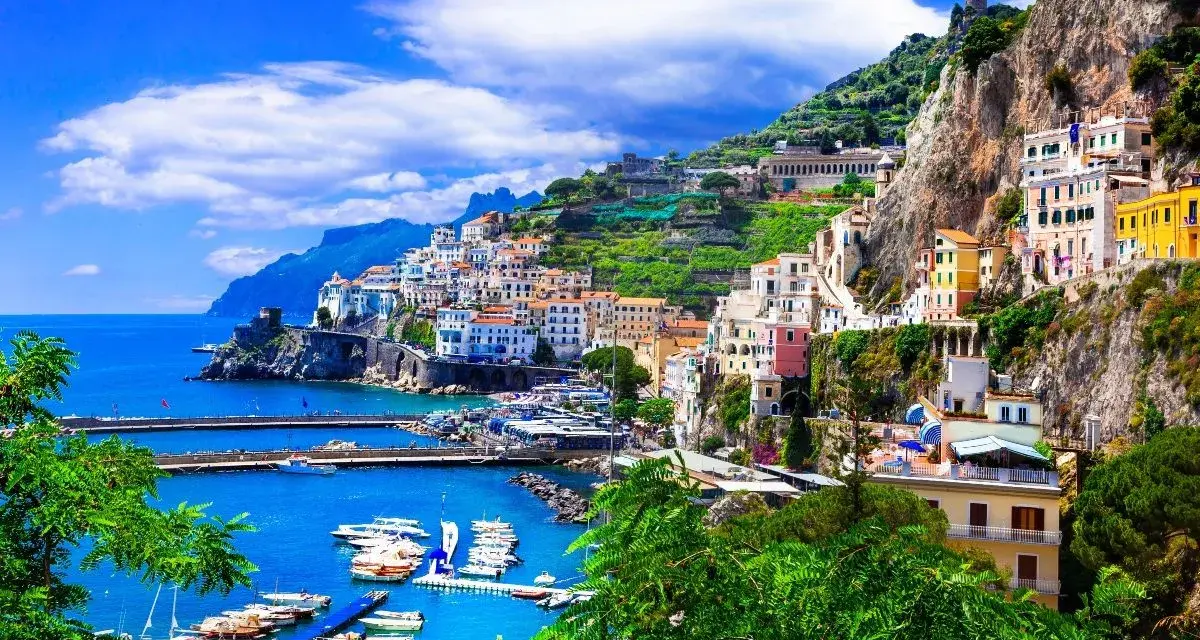 Un pueblo costero italiano con casas coloridas en laderas verdes, un puerto con barcos y el mar azul. ¡Un destino perfecto donde viajar en junio!