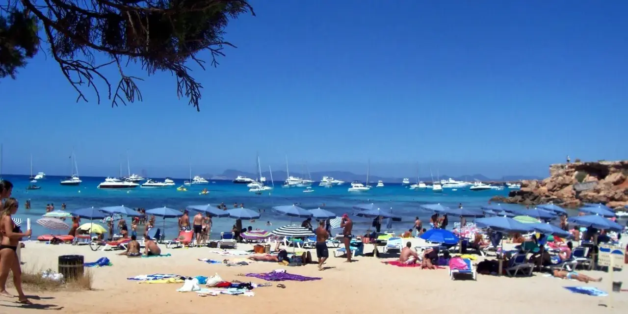 Playa con gente, sombrillas y barcos en el mar azul. Un día perfecto para ver qué ver en Formentera.