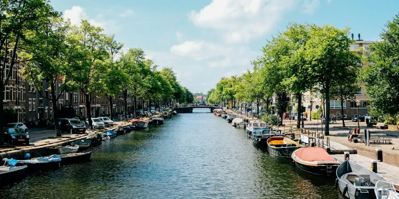 Canal de Ámsterdam con barcos, árboles y edificios bajo un cielo azul con nubes.