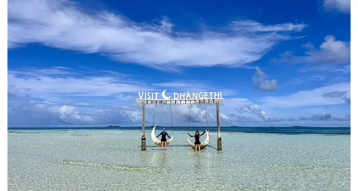 Dos personas disfrutan de columpios en forma de luna sobre aguas cristalinas. Un cartel dice "Visit Dhangethi", mostrando que las Maldivas low cost son posibles.