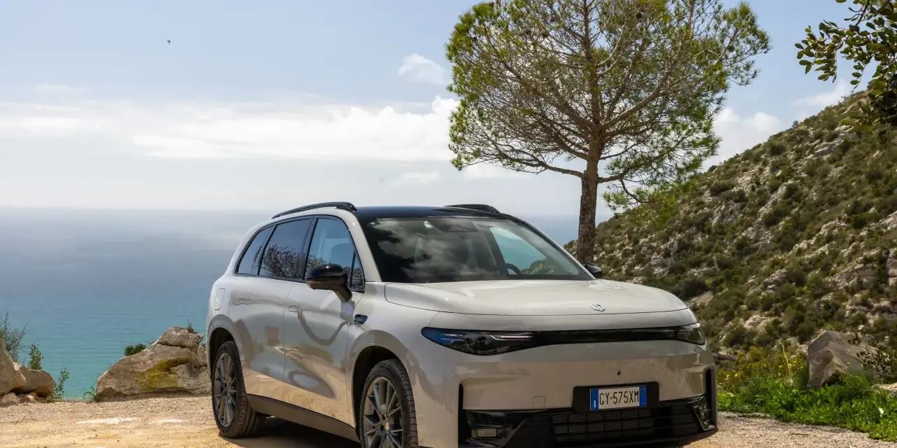 Un SUV blanco moderno, uno de los tipos de coches eléctricos, aparcado en un camino de tierra con el mar y una colina verde de fondo.