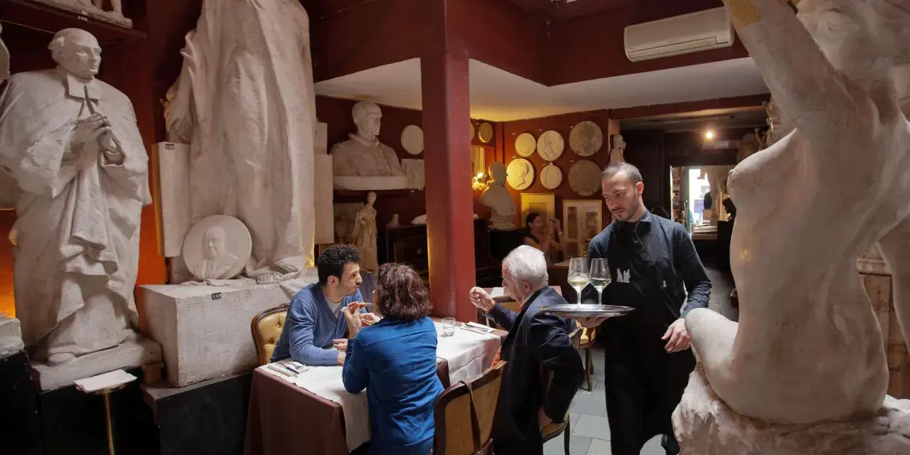Un camarero sirve copas de vino en un restaurante único, rodeado de esculturas. Un lugar ideal donde comer en Roma.