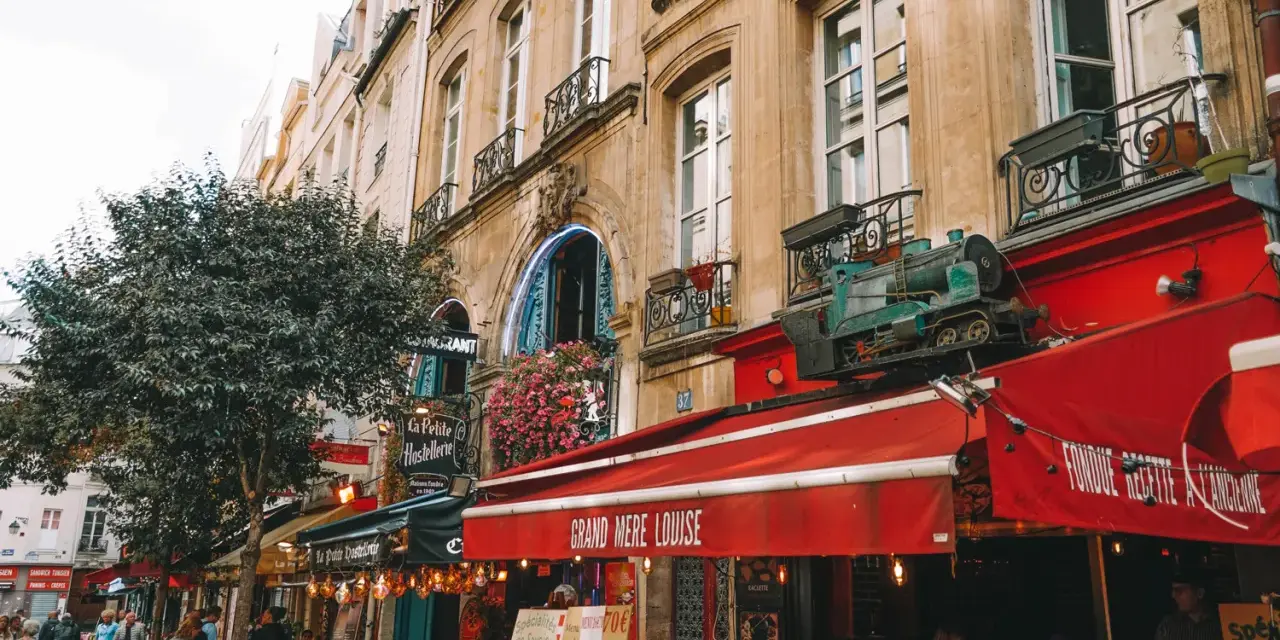 Calle parisina con restaurantes y mesas al aire libre, ideal para comer barato en París. Un tren decorativo adorna un toldo rojo.