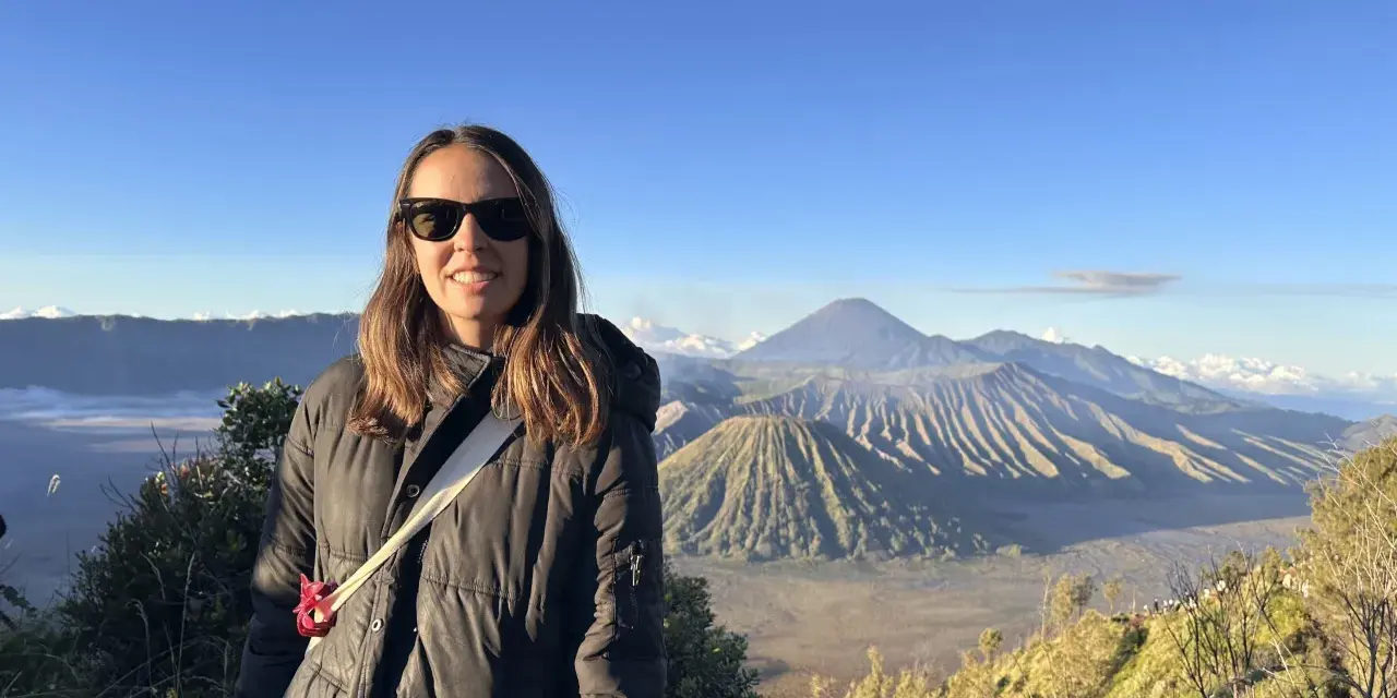 Mujer sonríe con gafas de sol frente a un paisaje volcánico en su ruta por Indonesia.