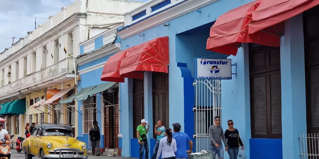 Gente paseando por una calle cubana, con un coche clásico amarillo y edificios de colores. Ideal para viajar a Cuba por libre.