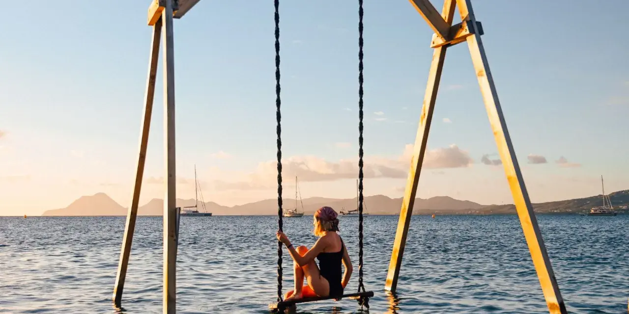 Mujer en columpio sobre el mar, ideal para saber donde viajar en febrero. Paisaje idílico con veleros y montañas al atardecer.