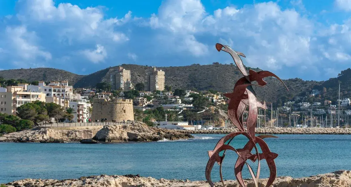 Escultura de aves metálicas en primer plano, con el castillo de Moraira y el puerto al fondo. Descubre qué ver en Moraira.