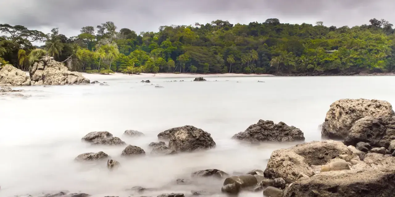 Costa Rica por libre: rocas y mar embravecido en una playa tropical, con densa vegetación al fondo.