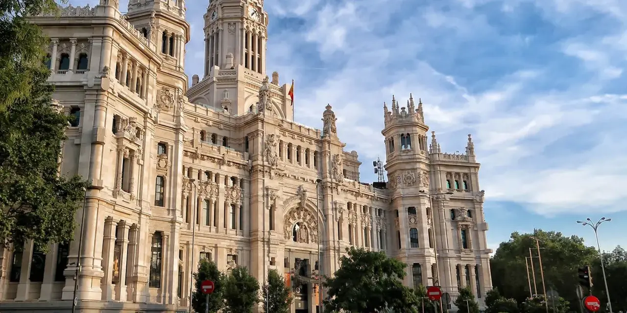 Un día en Madrid: el Palacio de Cibeles, un taxi blanco y coches circulan bajo un cielo azul con nubes.