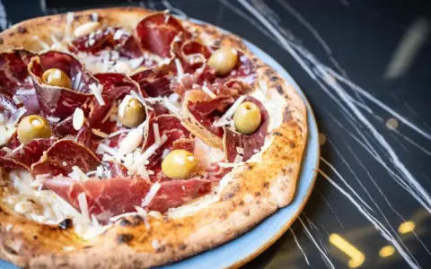 Pizza con jamón serrano, aceitunas y queso rallado, perfecta para saber donde comer en Menorca.