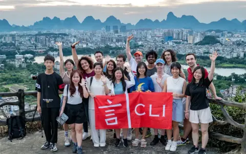 Grupo de jóvenes sonríe y saluda, sosteniendo una bandera roja con caracteres chinos y "CLI". Un paisaje urbano y montañoso de China al fondo, ideal para viajar a China por libre.