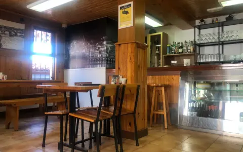 Mesas de madera y sillas listas para comer en Pontedeume. Barra con botellas y vasos, y un mural de la ciudad.