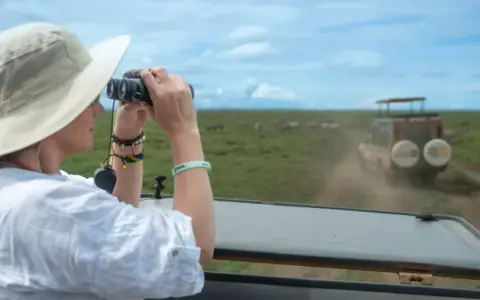 Mujer con sombrero y binoculares observa cebras en la sabana, disfrutando de su viaje a Tanzania.