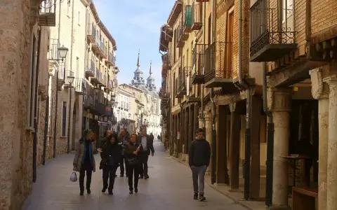 Gente pasea por una calle porticada, un lugar ideal para descubrir qué ver en Burgo de Osma. Edificios históricos y torres se alzan al fondo.