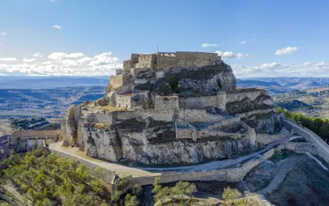 Castillo de Morella sobre un peñón rocoso, con murallas y edificios de piedra bajo un cielo azul con nubes.