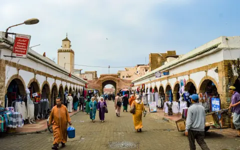 Mercado vibrante en Essaouira que ver: gente comprando ropa y artesanías bajo arcos blancos, con una mezquita al fondo.