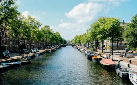 Canal de Ámsterdam con barcos, árboles y edificios bajo un cielo azul con nubes.