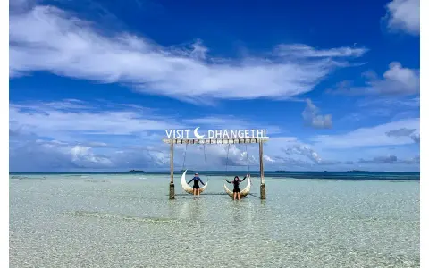 Dos personas disfrutan de columpios en forma de luna sobre aguas cristalinas. Un cartel dice "Visit Dhangethi", mostrando que las Maldivas low cost son posibles.
