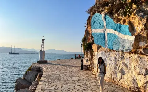 Una mujer posa junto a una roca con la bandera griega pintada. Un faro y un velero adornan la costa, ideal para saber qué ver en Grecia en una semana.