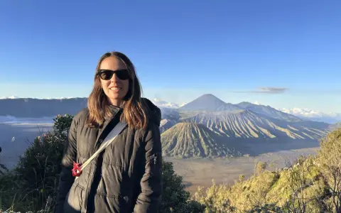 Mujer sonríe con gafas de sol frente a un paisaje volcánico en su ruta por Indonesia.