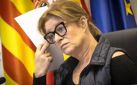 Mujer con gafas y chaleco oscuro, con banderas de España y Europa al fondo. Planeando su próximo viaje por España.