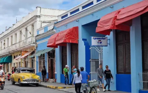 Gente paseando por una calle cubana, con un coche clásico amarillo y edificios de colores. Ideal para viajar a Cuba por libre.