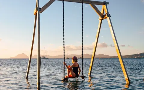 Mujer en columpio sobre el mar, ideal para saber donde viajar en febrero. Paisaje idílico con veleros y montañas al atardecer.