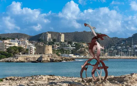 Escultura de aves metálicas en primer plano, con el castillo de Moraira y el puerto al fondo. Descubre qué ver en Moraira.