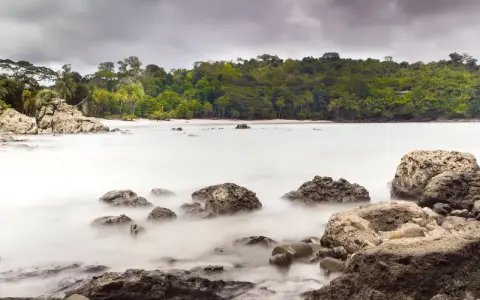 Costa Rica por libre: rocas y mar embravecido en una playa tropical, con densa vegetación al fondo.