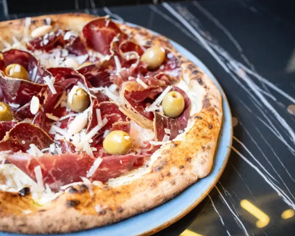 Pizza con jamón serrano, aceitunas y queso rallado, perfecta para saber donde comer en Menorca.