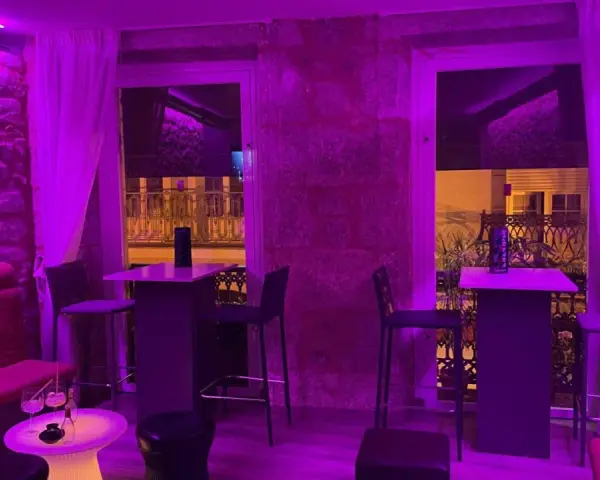Ambiente moderno y acogedor para comer en Coruña. Mesas altas, taburetes y sofás rojos bajo luces violetas.